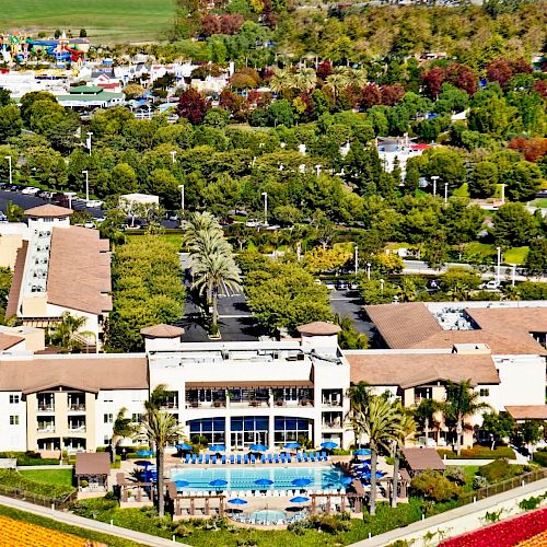 Aerial view of a resort with a large hotel, pool, palm trees, landscaped grounds, and colorful flower beds in the foreground and surrounding greenery.