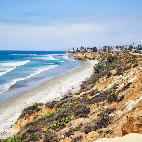 Crescent beach with gentle waves, a sandy shore, and rugged cliffs; distant houses line the horizon under a clear, blue sky.