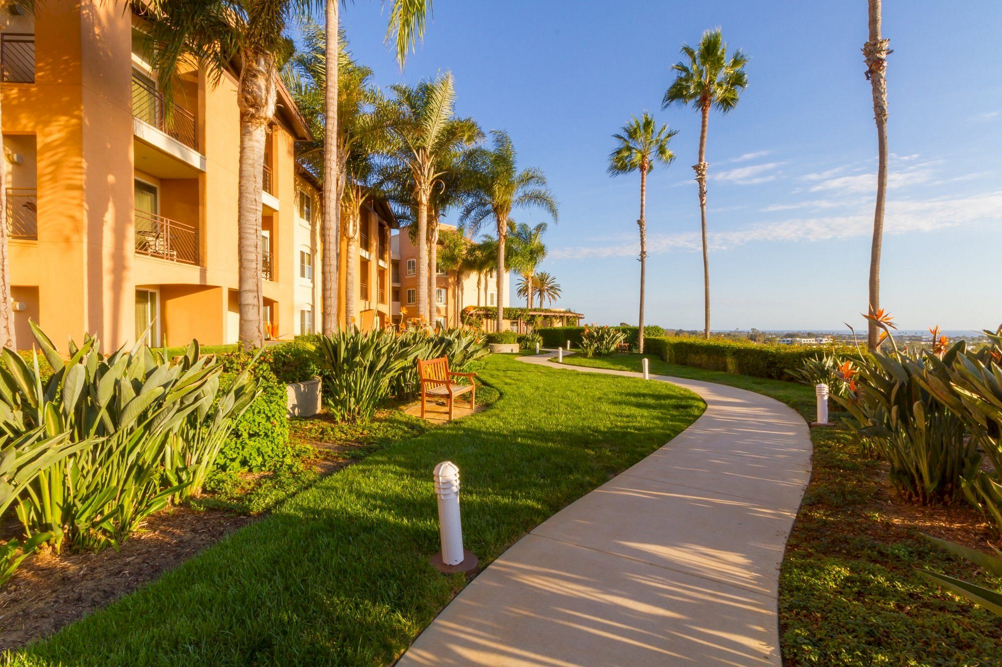 A sunny resort path flanked by palm trees and green lawns winds past a tan building toward the ocean, inviting a peaceful seaside stroll.