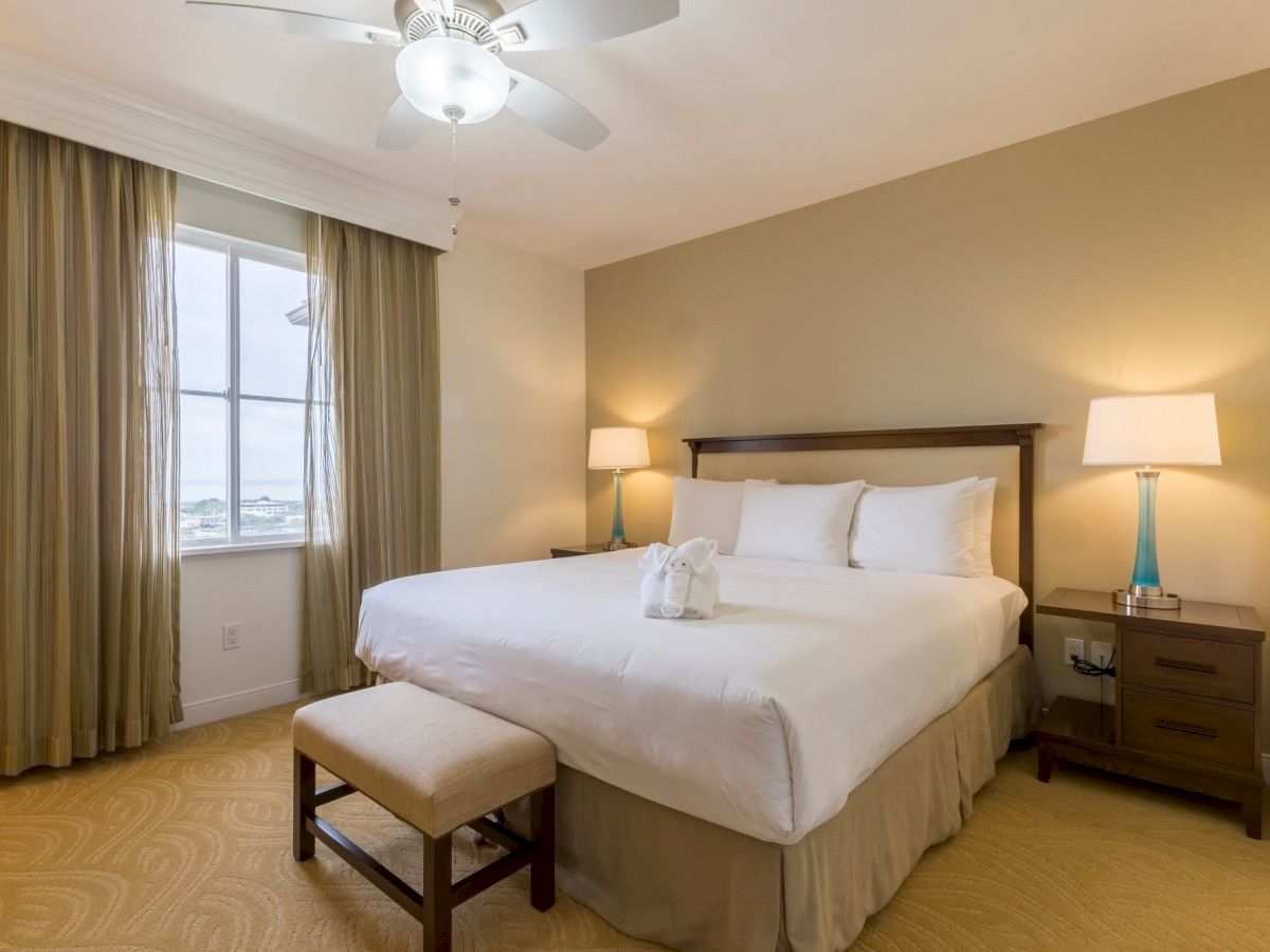 A cozy hotel bedroom with a king bed, two nightstands with lamps, a cushioned bench, beige walls, and soft natural light from a window with long curtains.