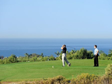 Two people on a sunny golf course overlooking the sea, one swinging a club while the other watches, with green grass and sparse shrubs in the foreground.