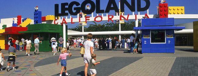 A sunny entrance to LEGOLAND California with colorful toy-block structures, families walking toward the gates, and a large LEGO sign above.
